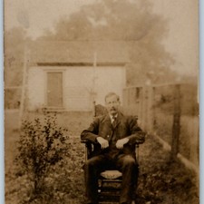 c1910s RPPC Man Seated in Wooden Armchair Outdoors Yard Shed Fence Suit Portrait picture